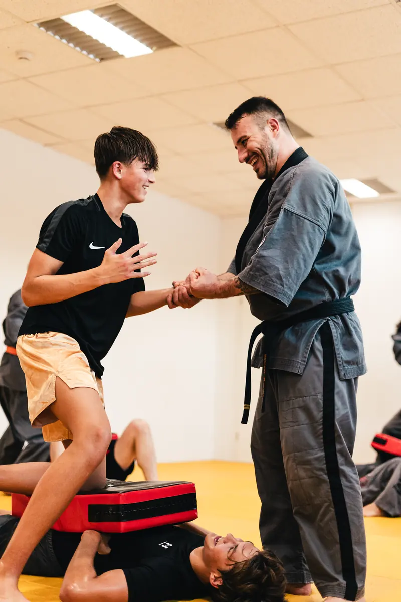 Ein Trainer im Gi und ein Jugendlicher reichen sich lachend die Hand auf der Matte
