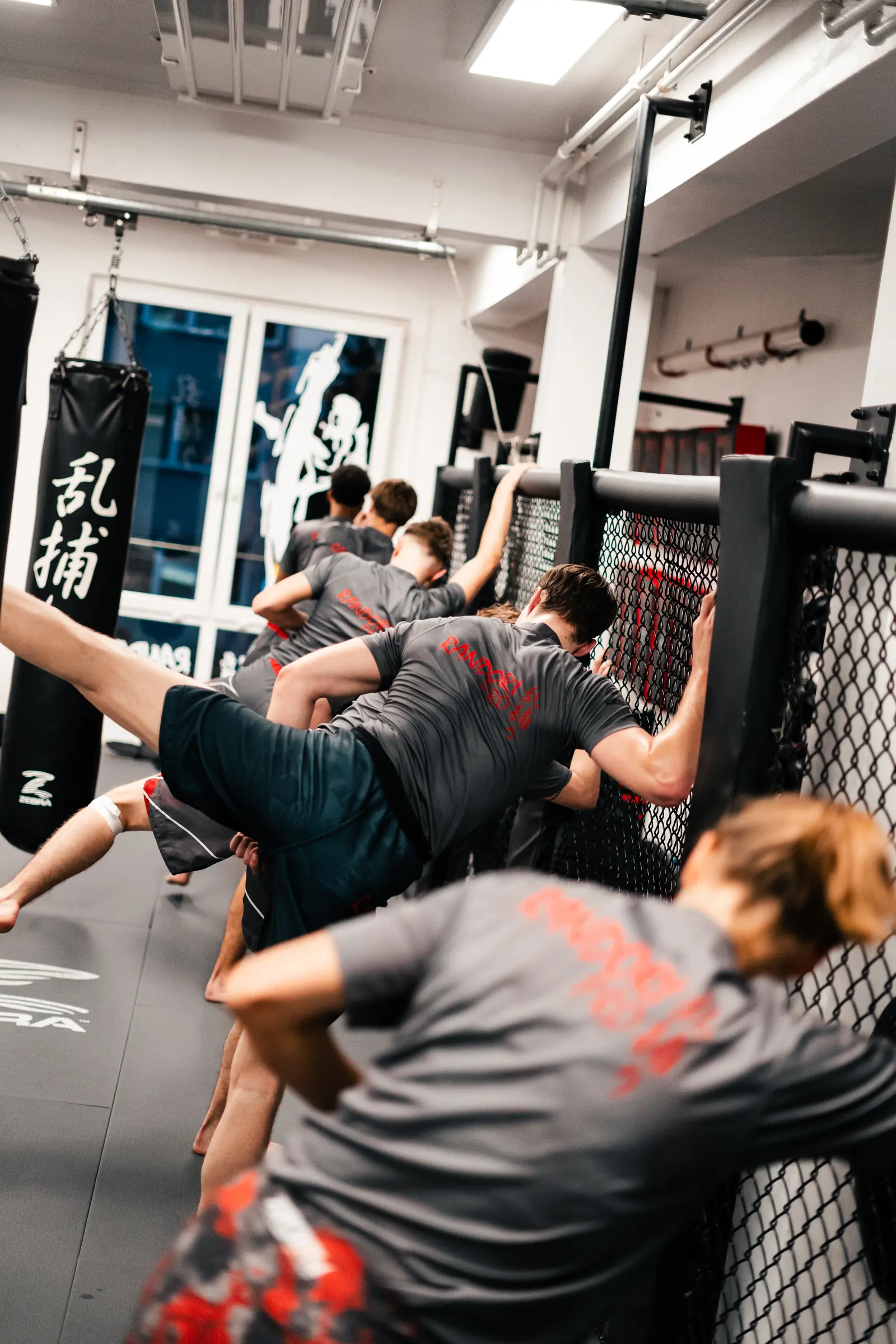 Trainingsgruppe in Randori-Pro-Shirts trainiert Tritte, an den Käfigzaun gestützt