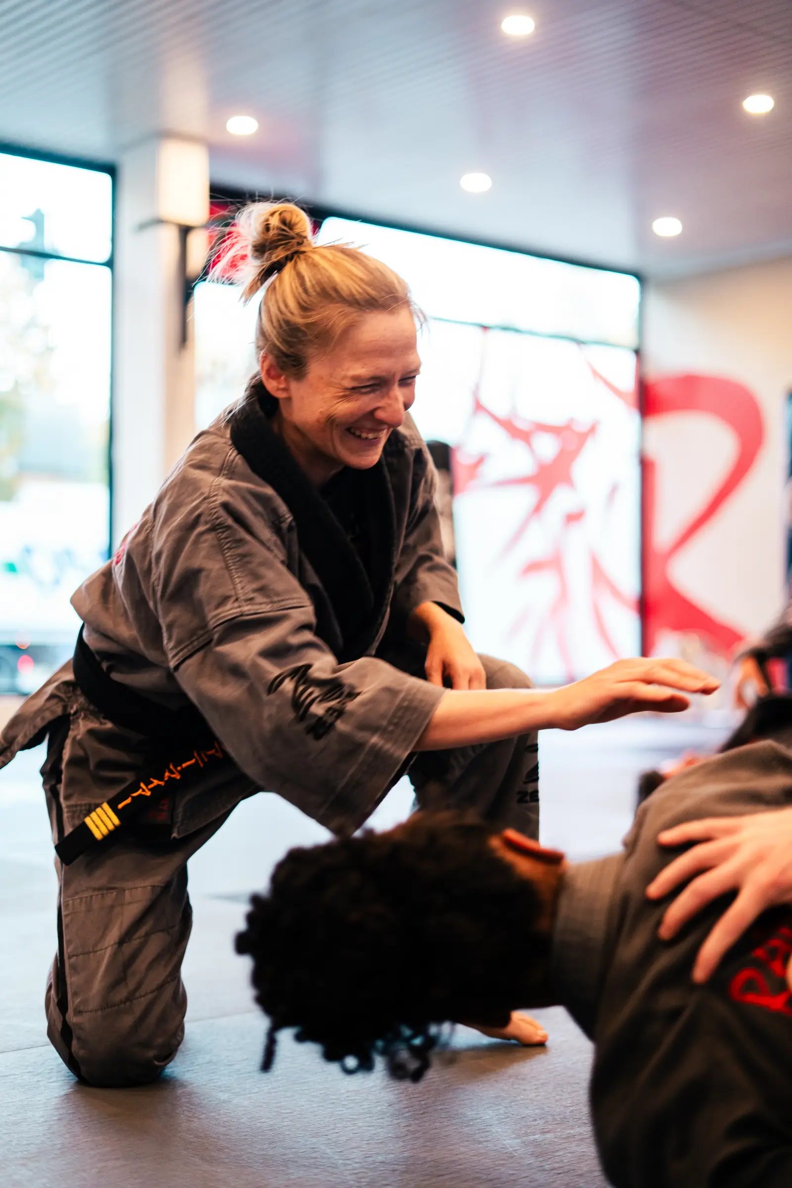 Eine Frau im Gi kniet lachend auf der Matte und übt mit einem Trainingspartner