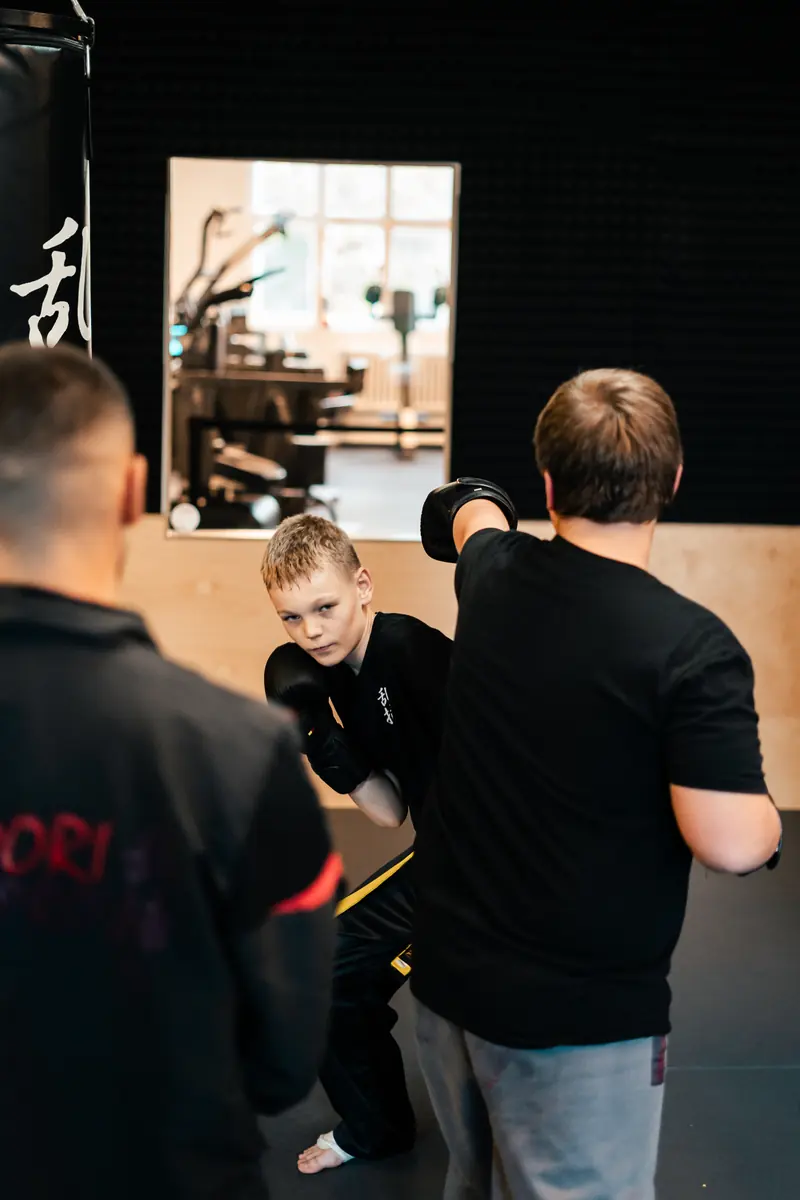 Ein Junge mit Boxhandschuhen schlägt auf die Pratze seines Gegenübers, im Vordergrund schaut ein Trainer zu